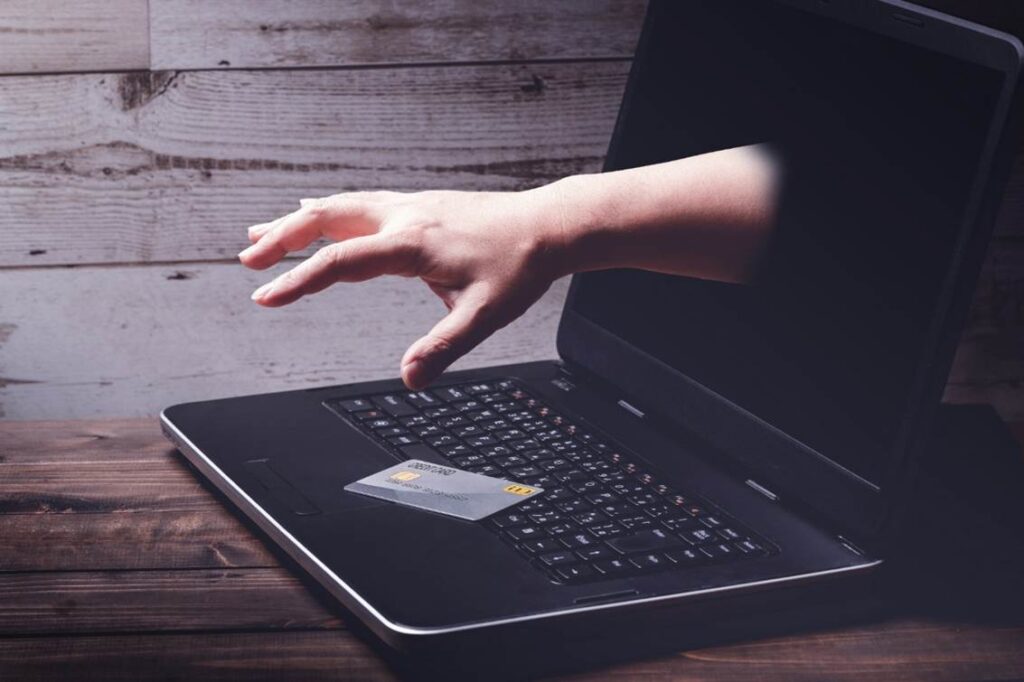 キーボード上に置かれたクレジットカードを掴み取ろうとする、パソコンの画面から伸びる手